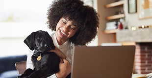 Smiling young women of colour holds her black labrador puppy in front of her open laptop