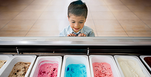 A young boy happily looking at seven different flavours of ice-cream through a protective screen