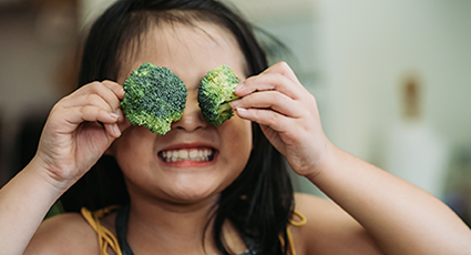 Young girl smiling whilst holding two broccoli florets up to her eyes.