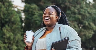 Young black woman walking and laughing whilst holding a coffee cup and a laptop