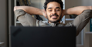 Young man sitting down smiling in front of laptop with hands behind his head relaxing