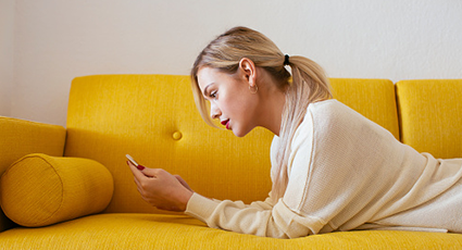 Young woman lying on a yellow sofa, using her phone 425x230
