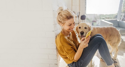Young woman sitting down with her dog, whilst smiling and looking at her mobile phone