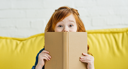 Young wide-eyed girl with red hair, peeking over a book