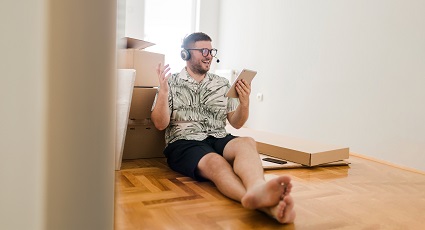 Young white male sitting on floor next to some cardboard boxes, wearing a headset, whilst smiling at his tablet device
