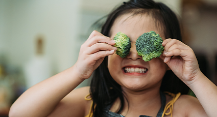 Child smiling and holding broccoli florets over eyes